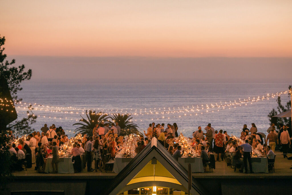 A large outdoor gathering at sunset near the ocean, with string lights overhead and tables full of people dining.