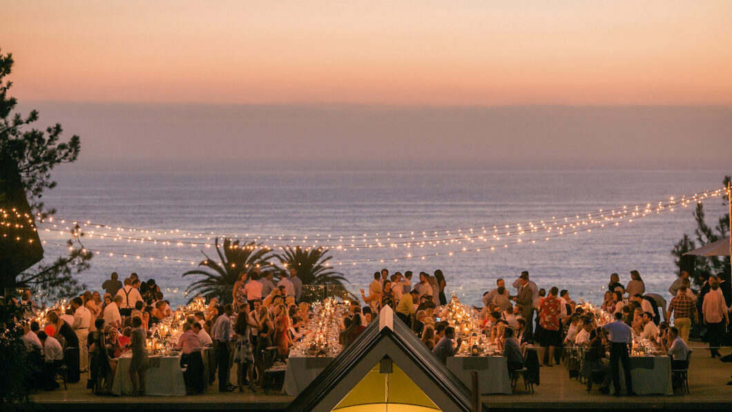 A large outdoor gathering at sunset near the ocean, with string lights overhead and tables full of people dining.