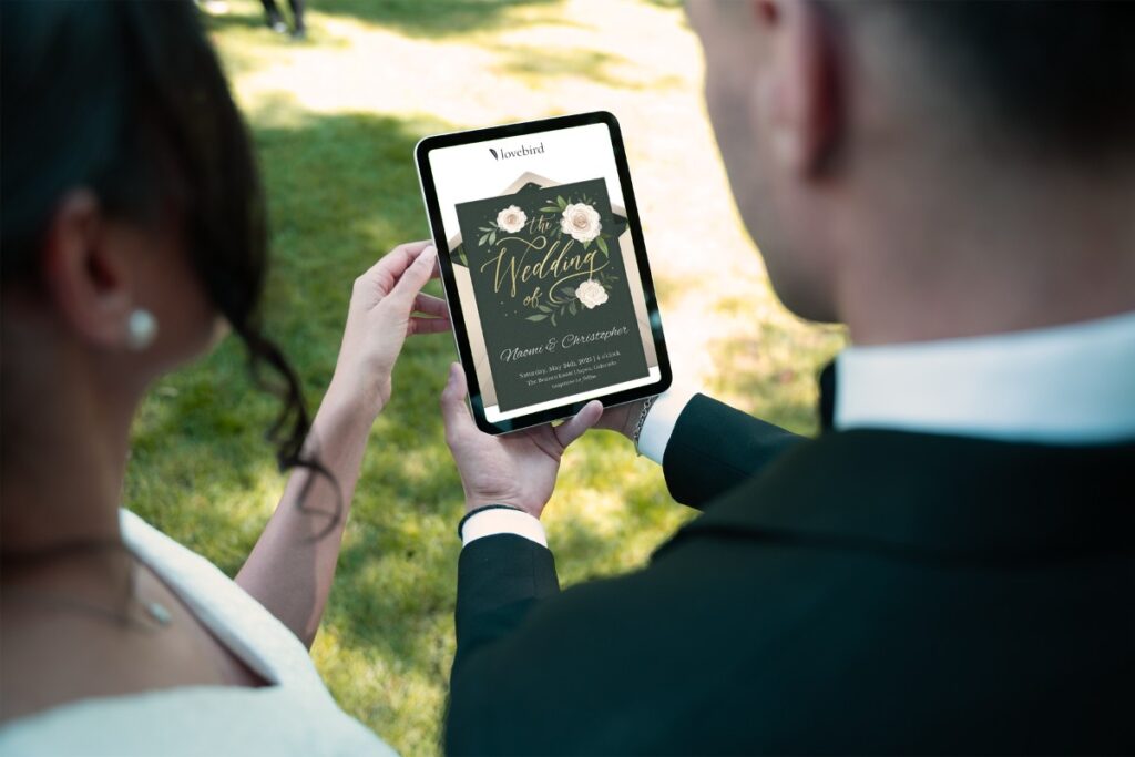 A couple holds a tablet displaying a digital wedding invitation with floral designs on a sunny day.