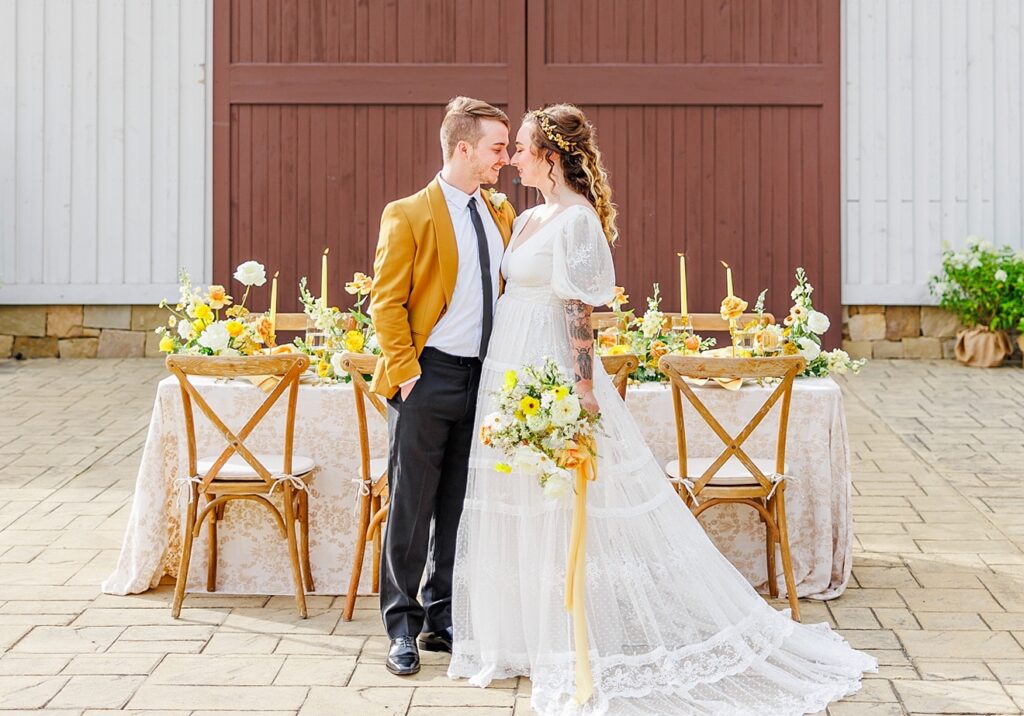 A couple stands in front of a decorated wedding table. The man wears a mustard jacket; the woman is in a white lace dress holding a bouquet. The backdrop features a wooden barn door.