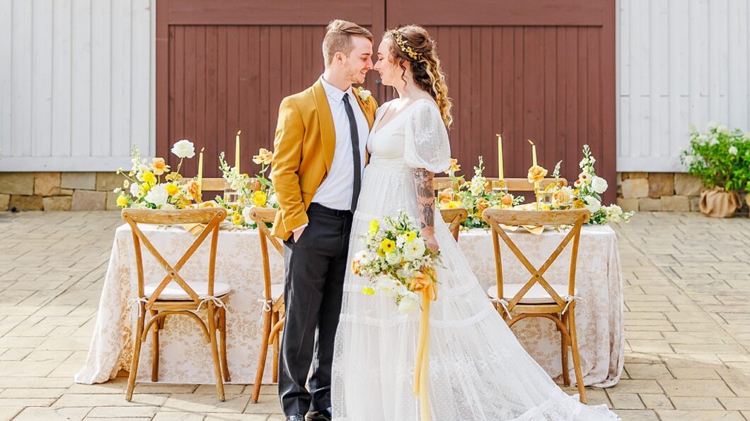 A couple stands in front of a decorated wedding table. The man wears a mustard jacket; the woman is in a white lace dress holding a bouquet. The backdrop features a wooden barn door.