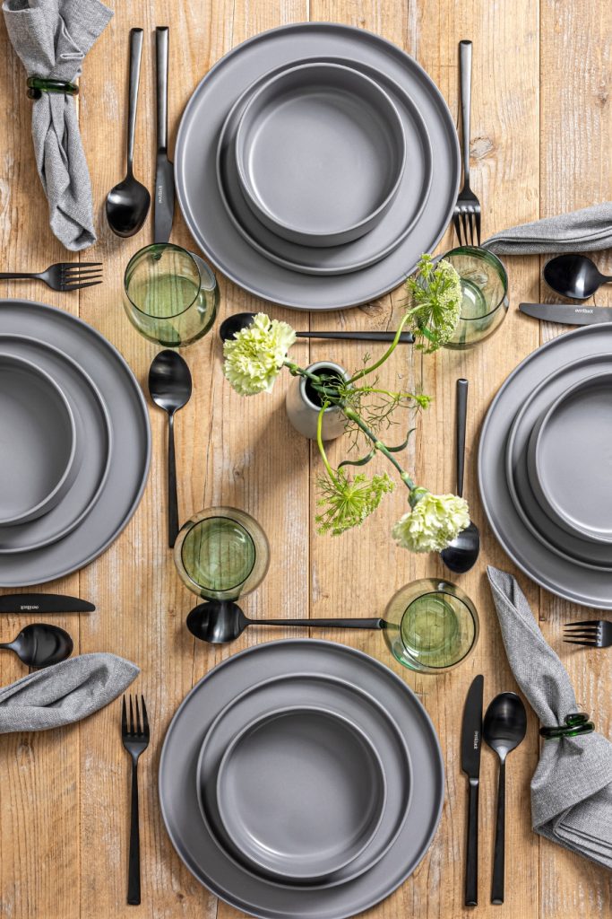 A wooden table set for four with gray plates, black cutlery, green glasses, gray napkins, and a small flower vase in the center.