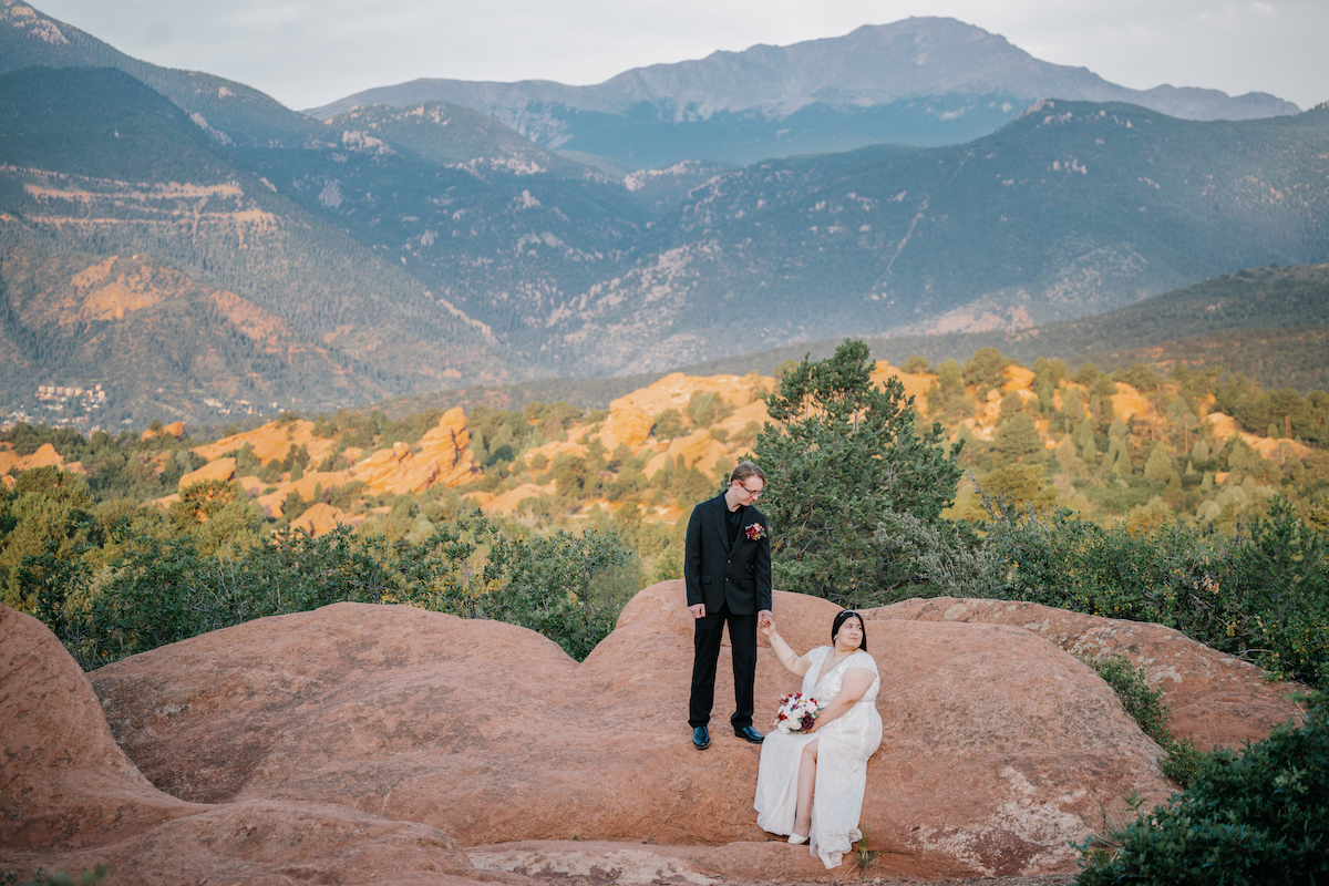 A couple dressed in formal attire stands on rocky terrain with green foliage and mountains in the background.