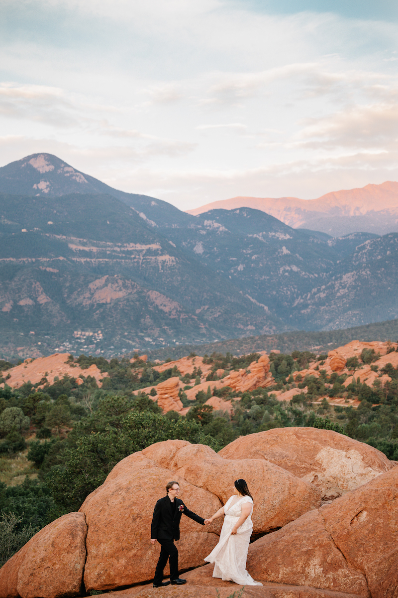 A couple stands on large red rocks, holding hands, with green hills and distant mountains in the background under a partly cloudy sky.