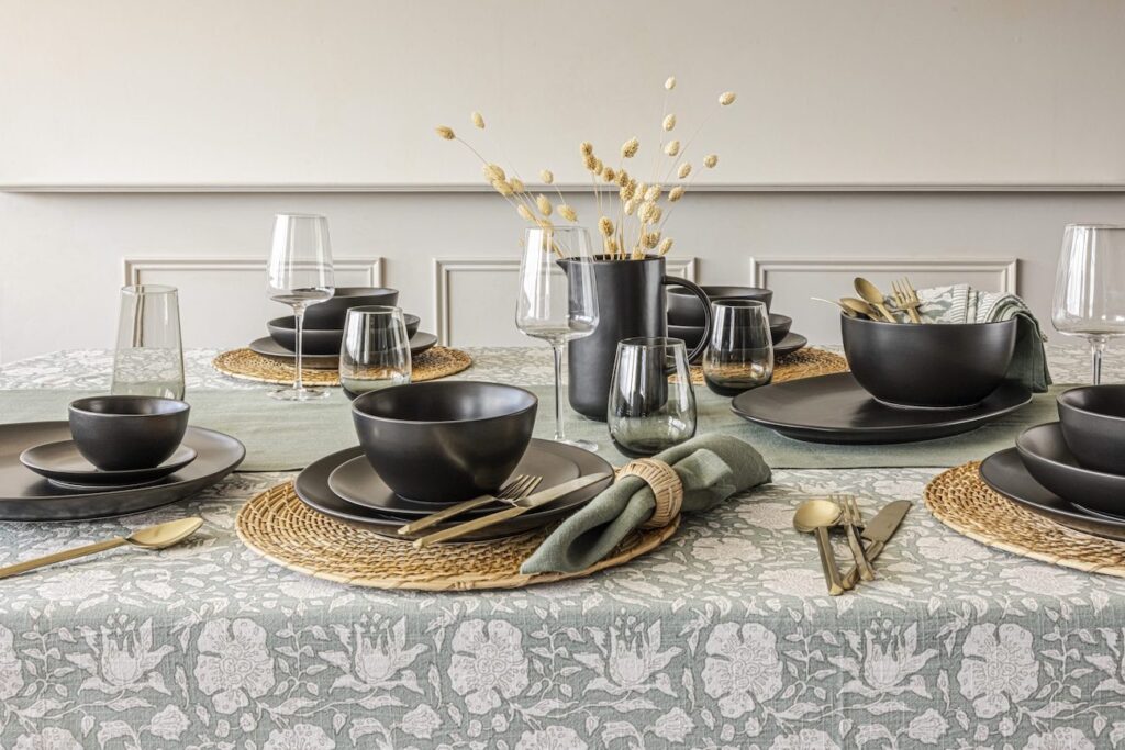 A dining table is set with black dinnerware, gold cutlery, and glasses on a patterned tablecloth. A black vase with dried flowers is in the center.