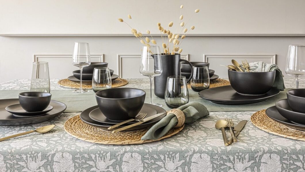 A dining table is set with black dinnerware, gold cutlery, and glasses on a patterned tablecloth. A black vase with dried flowers is in the center.