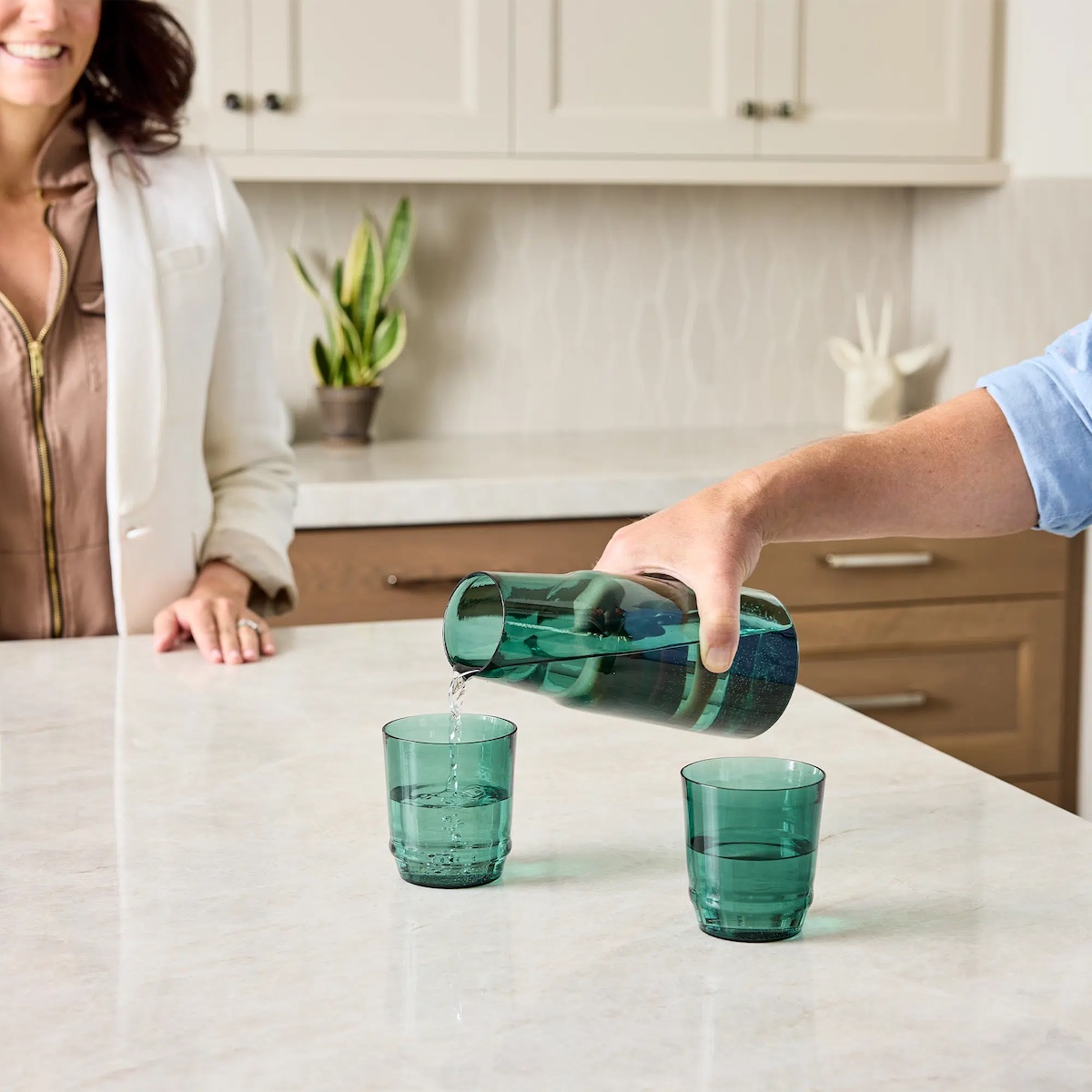 A person pours water from a green pitcher into a green glass cup on a white kitchen countertop, with another person standing nearby.