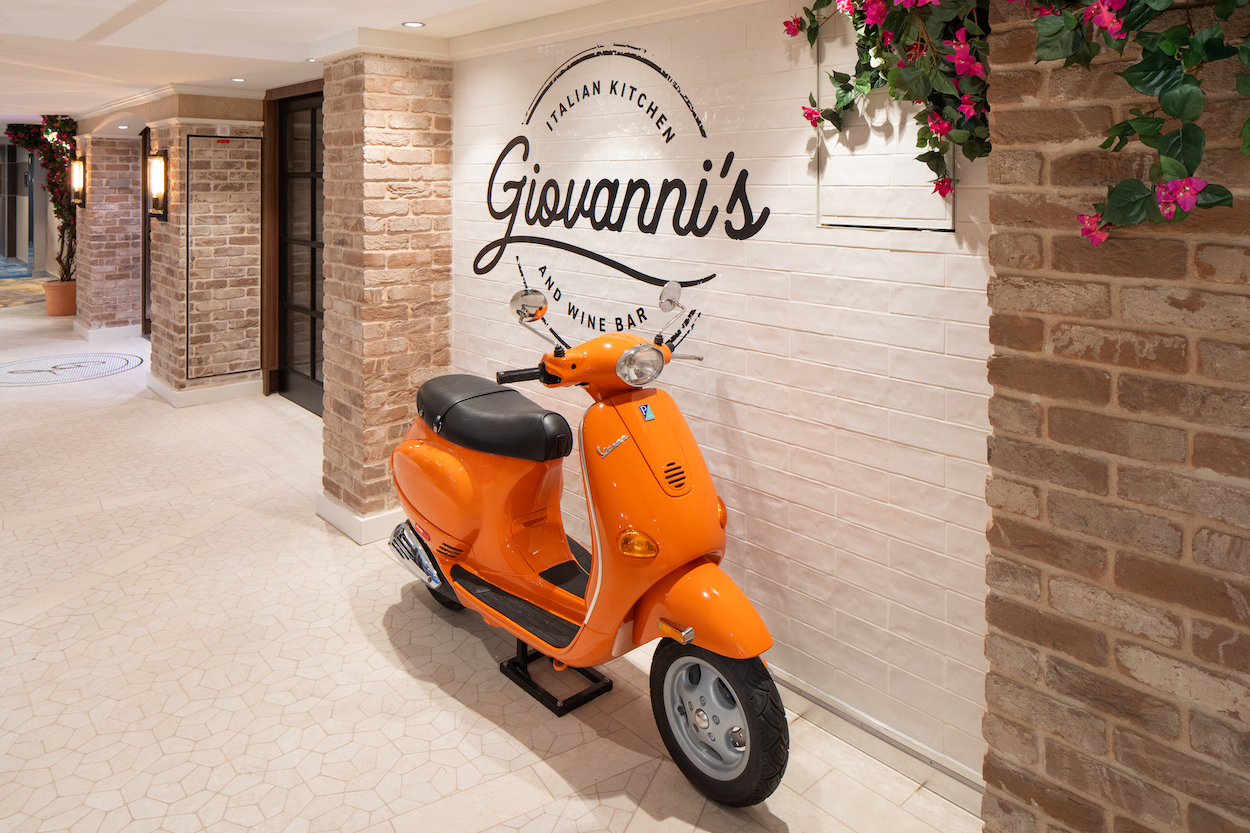 Orange scooter parked against a brick wall with "Giovanni's Italian Kitchen &amp; Wine Bar" sign, and flowers decorate the top.