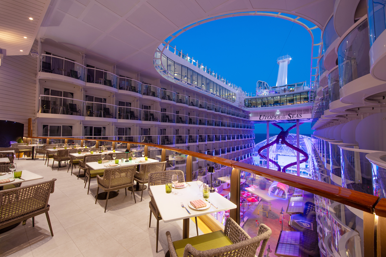 A spacious cruise ship interior with balconies, outdoor dining tables, and a view of the sky and upper deck structures.