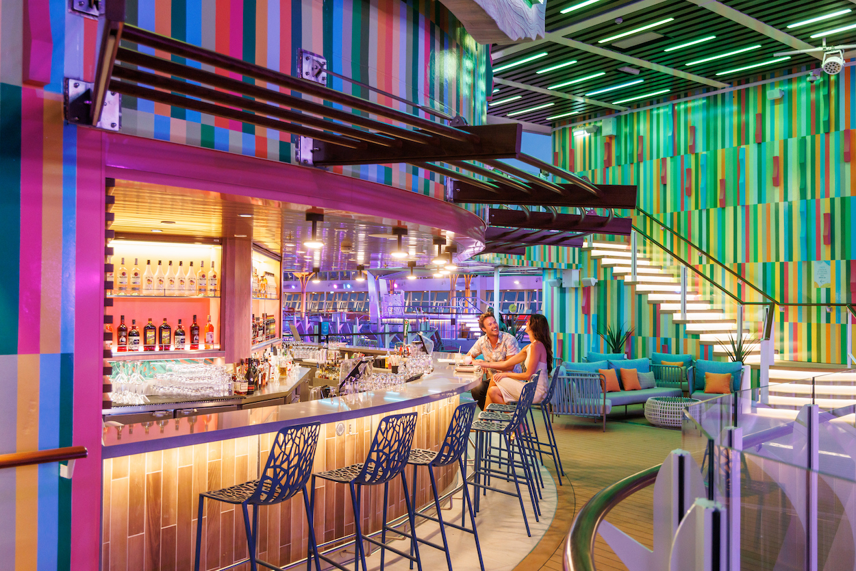 A vibrant bar with colorful striped walls and neon lighting. Several people sit at the bar on high stools, and others relax on couches. A staircase leads to an upper level.