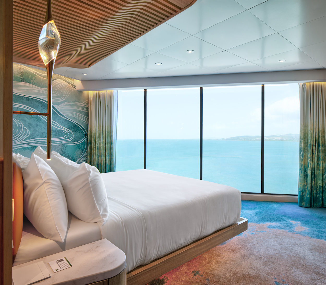 Modern hotel room with a large bed, decorative ceiling, abstract wall art, and expansive windows offering an ocean view.