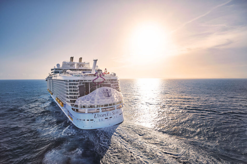 A large cruise ship sails on the ocean under a clear sky with the sun setting on the horizon.