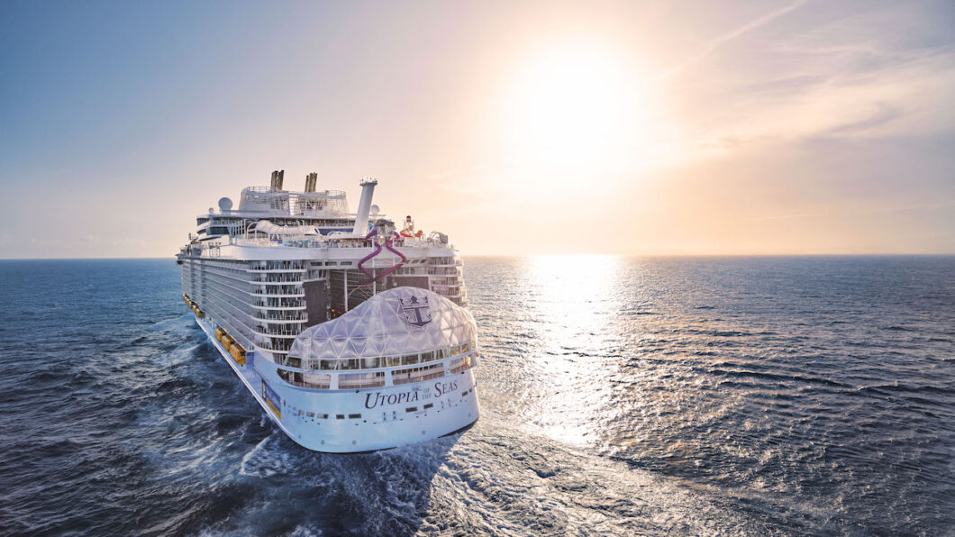 A large cruise ship sails on the ocean under a clear sky with the sun setting on the horizon.