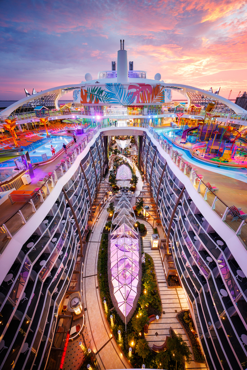 Aerial view of a cruise ship's top deck featuring pools, colorful play areas, and a central garden with glass-covered pathways during sunset.