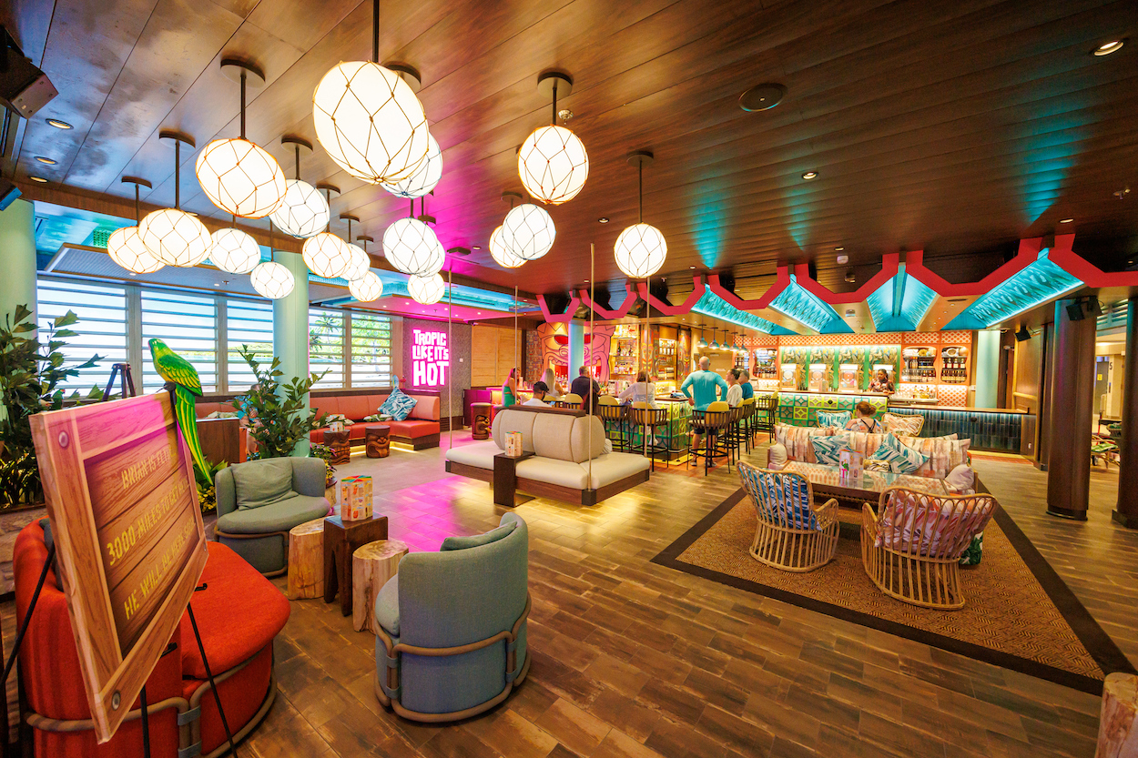 A colorful tropical-themed lounge with round hanging lights, vibrant decor, seating areas, and a bar in the background. A neon sign reads "Tropic Like It's Hot.