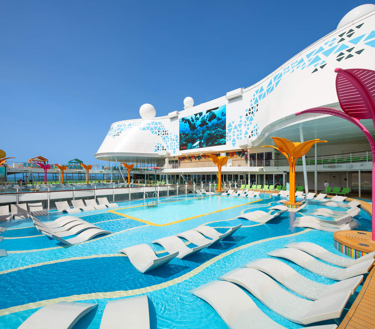 A cruise ship deck with a bright blue pool, surrounded by lounge chairs and colorful decorative structures under a clear blue sky.