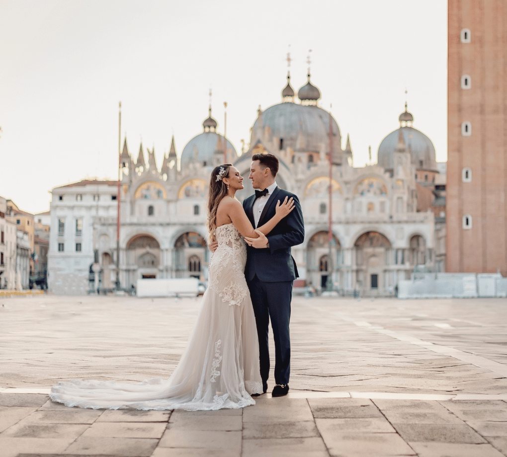 Bride and groom stand together in front of a historic building with domes and arches. The bride wears a long white gown, and the groom is in a dark suit.