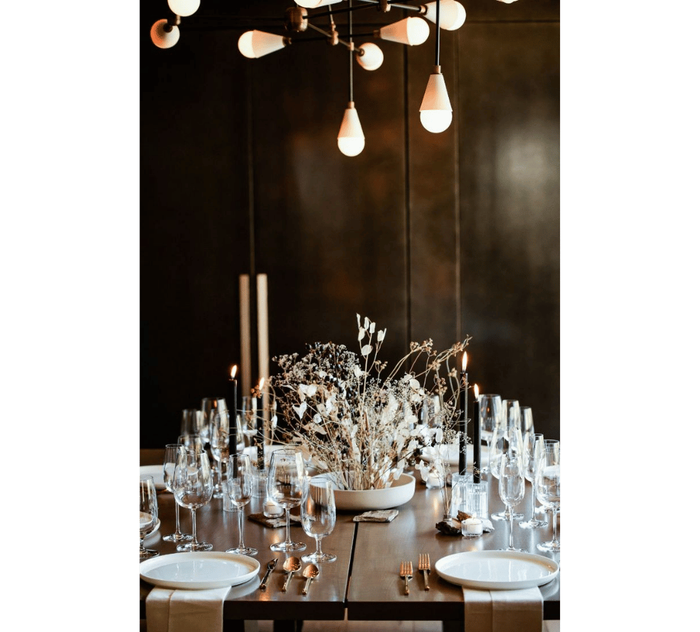 Elegant dining table set with white plates, glassware, and cutlery. A centerpiece of dried flowers and candles under a modern chandelier with hanging bulbs creates a warm ambiance.