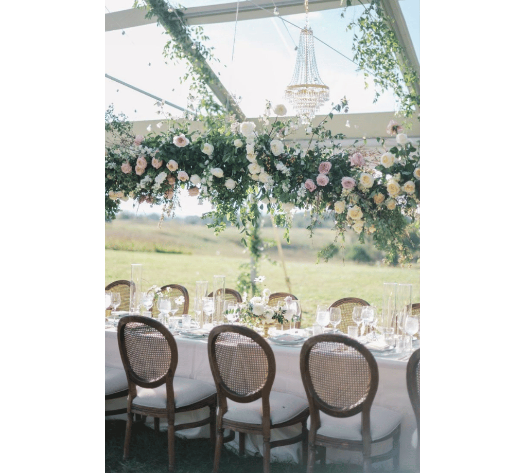 A long table set for an elegant outdoor event with wooden chairs and white tablecloth. Floral arrangements and a chandelier hang overhead, with a scenic view visible through clear tent walls.