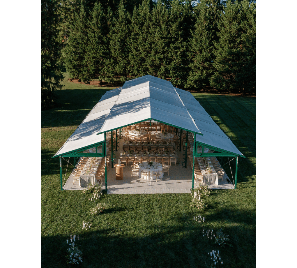 Outdoor tent setup with rows of tables and chairs on a grassy area, surrounded by trees.