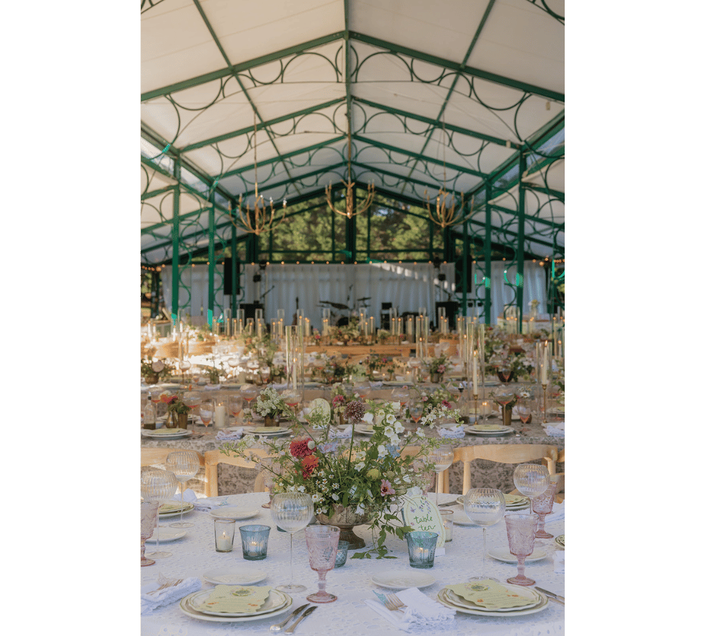 Elegant outdoor venue set for an event. Tables are adorned with floral centerpieces, glassware, and cutlery under a green and white canopy.