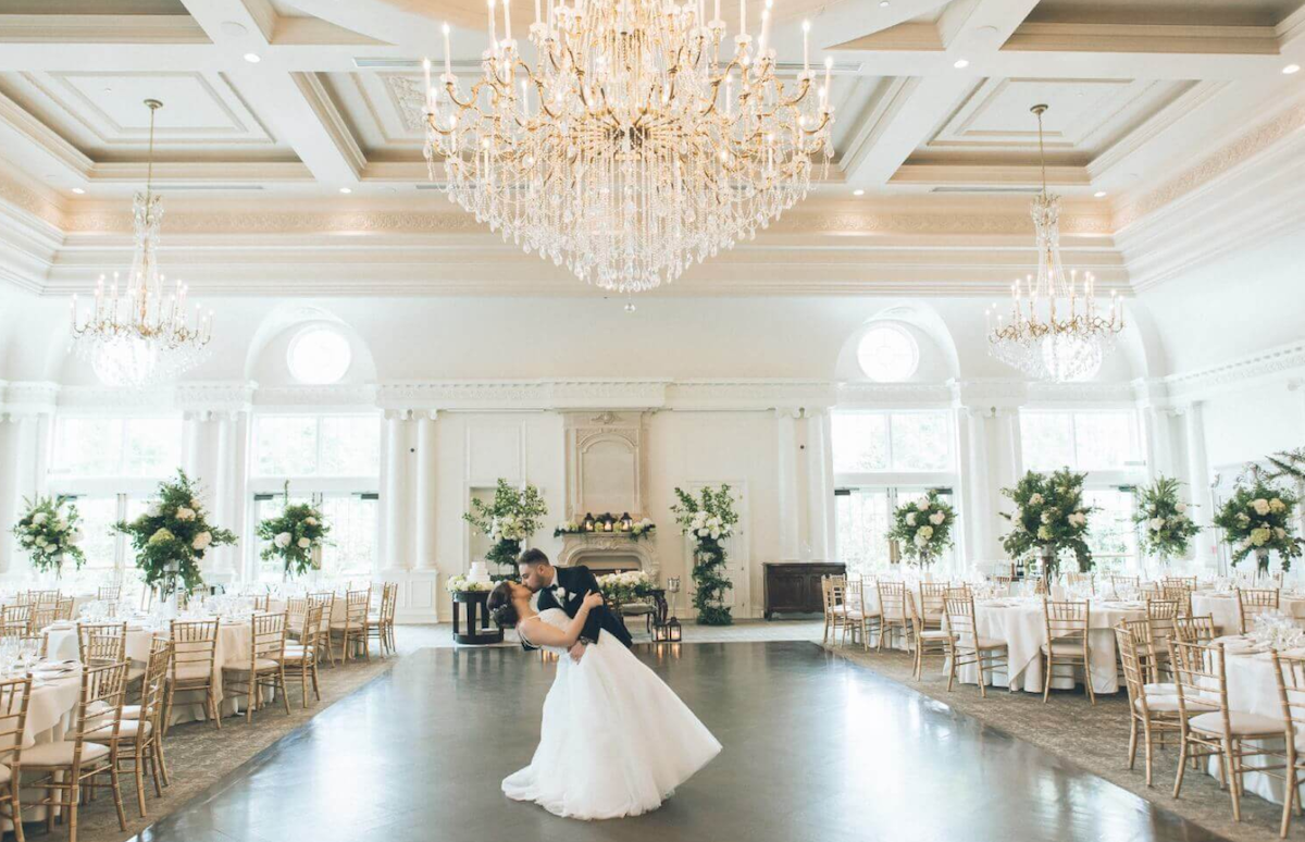 A couple dances in an elegant ballroom decorated with chandeliers, white walls, and floral arrangements. Tables with gold chairs surround the dance floor.