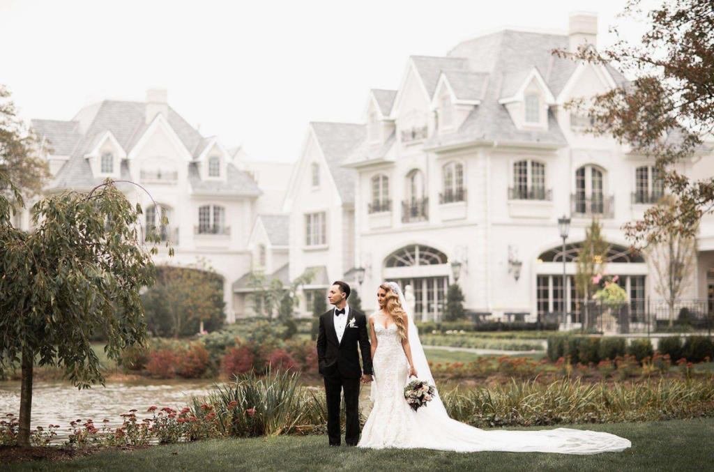 A bride and groom stand together in front of a large, elegant mansion with gardens and a pond. The groom wears a black suit, and the bride is in a white gown holding a bouquet.