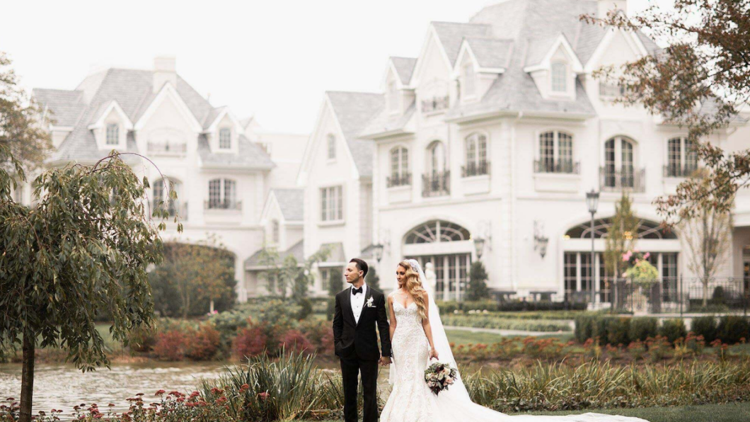 A bride and groom stand together in front of a large, elegant mansion with gardens and a pond. The groom wears a black suit, and the bride is in a white gown holding a bouquet.