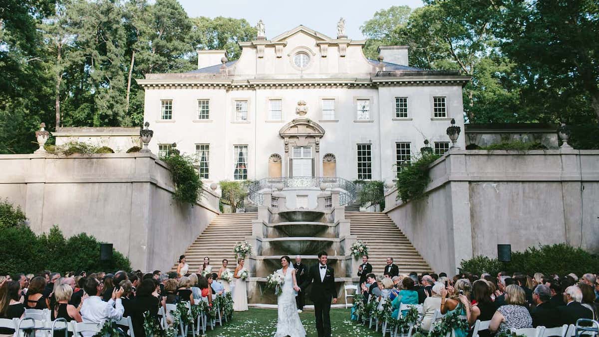 A bride and groom walk down the aisle outdoors, surrounded by seated guests. The setting includes a grand staircase and an elegant building in the background.