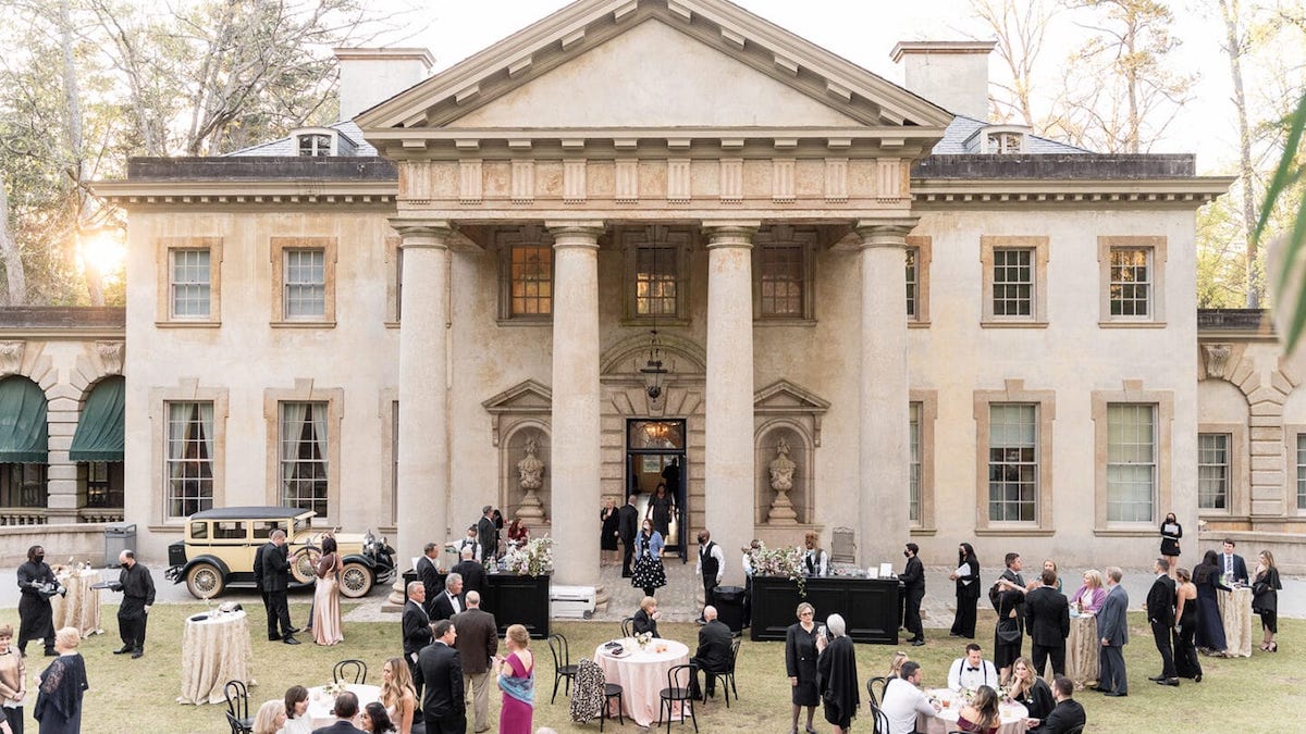 People in formal attire gather outside a grand mansion with pillars, surrounded by trees, and vintage car parked nearby.