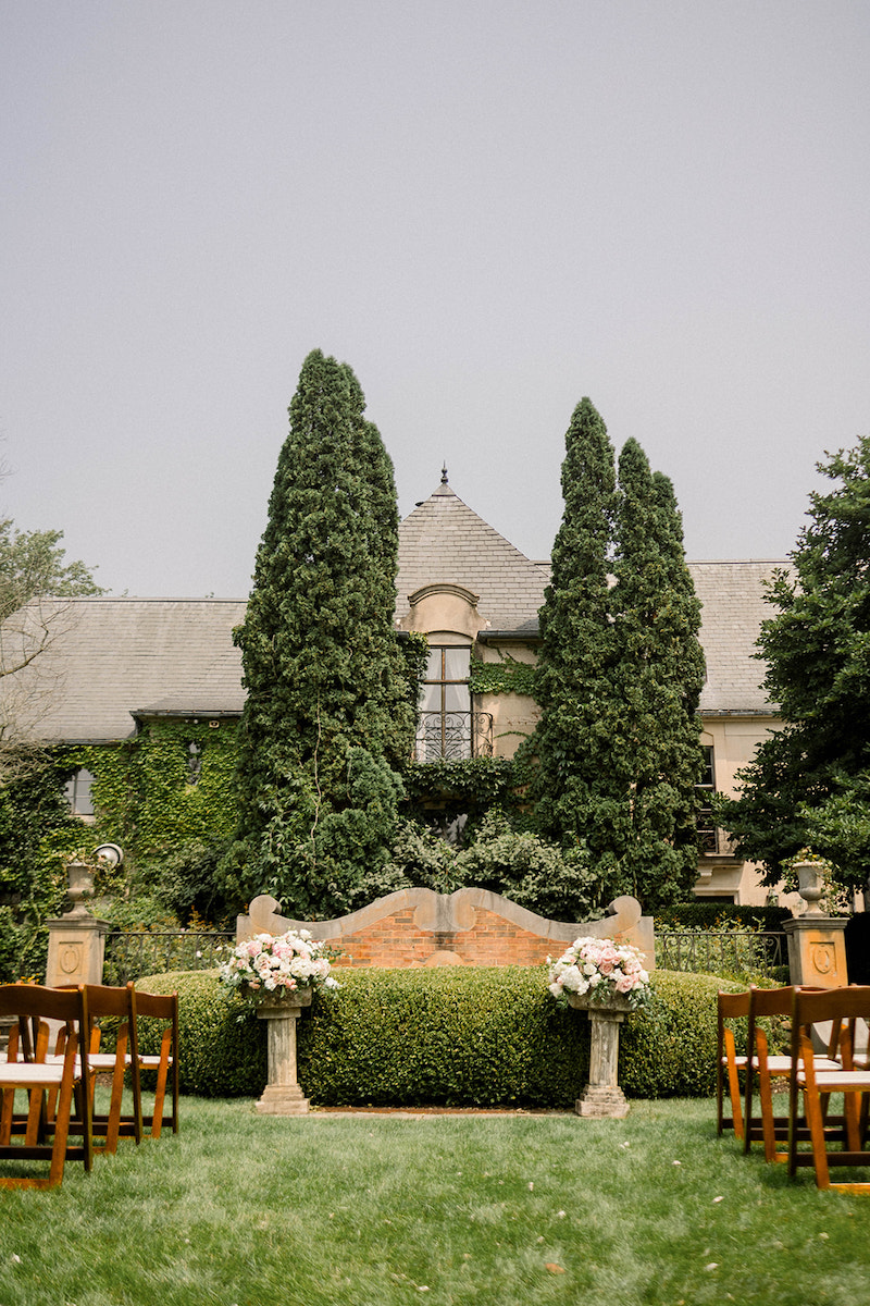 Outdoor wedding setup with wooden chairs facing a floral-decorated stone altar. Ivy-covered building and tall trees in the background.