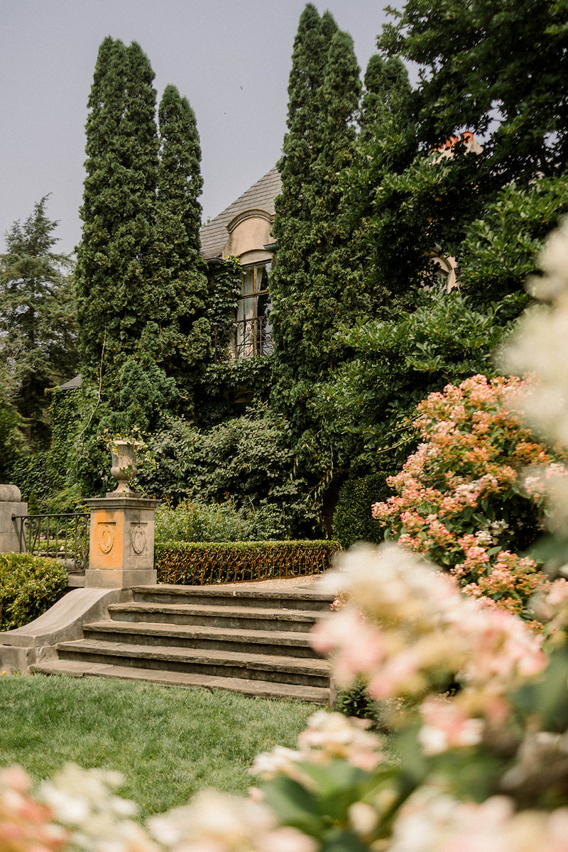 A stone building partially covered in ivy with tall trees and manicured gardens. A stone staircase leads to the entrance, surrounded by blooming flowers and green shrubs.