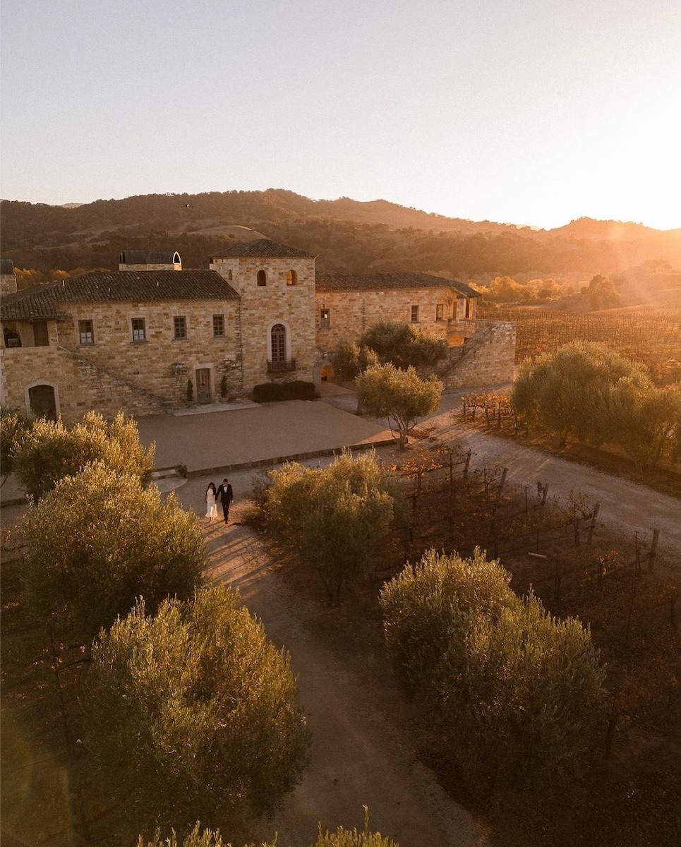 Aerial view of a couple walking near a rustic stone building surrounded by trees and vineyards at sunset.