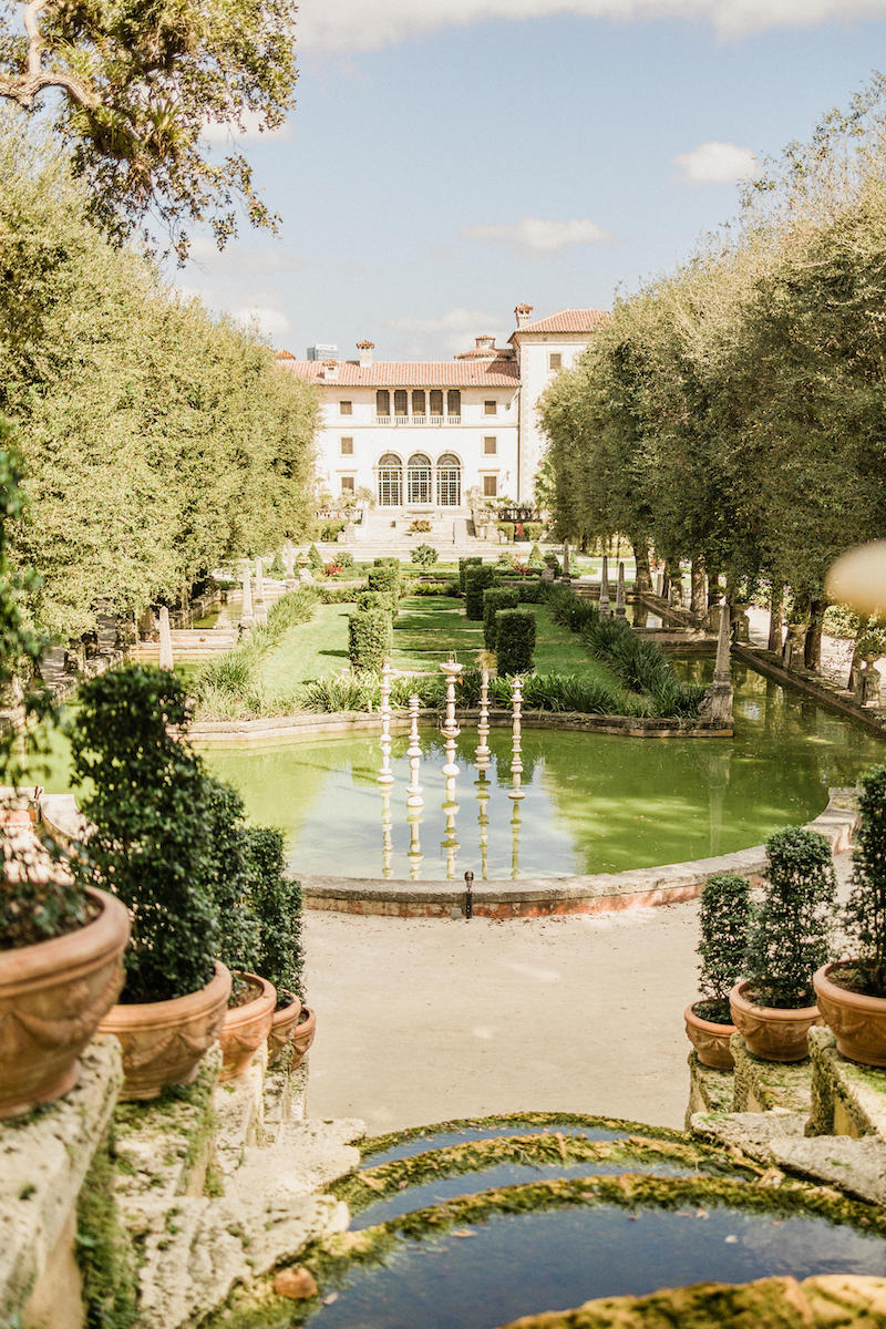 Elegant garden with trimmed hedges, a circular pond with fountains, and a historic villa in the background.