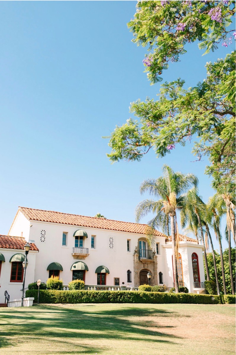 A two-story white building with red-tiled roof, arched windows, and green awnings is surrounded by palm trees and lush landscaping under a clear blue sky.