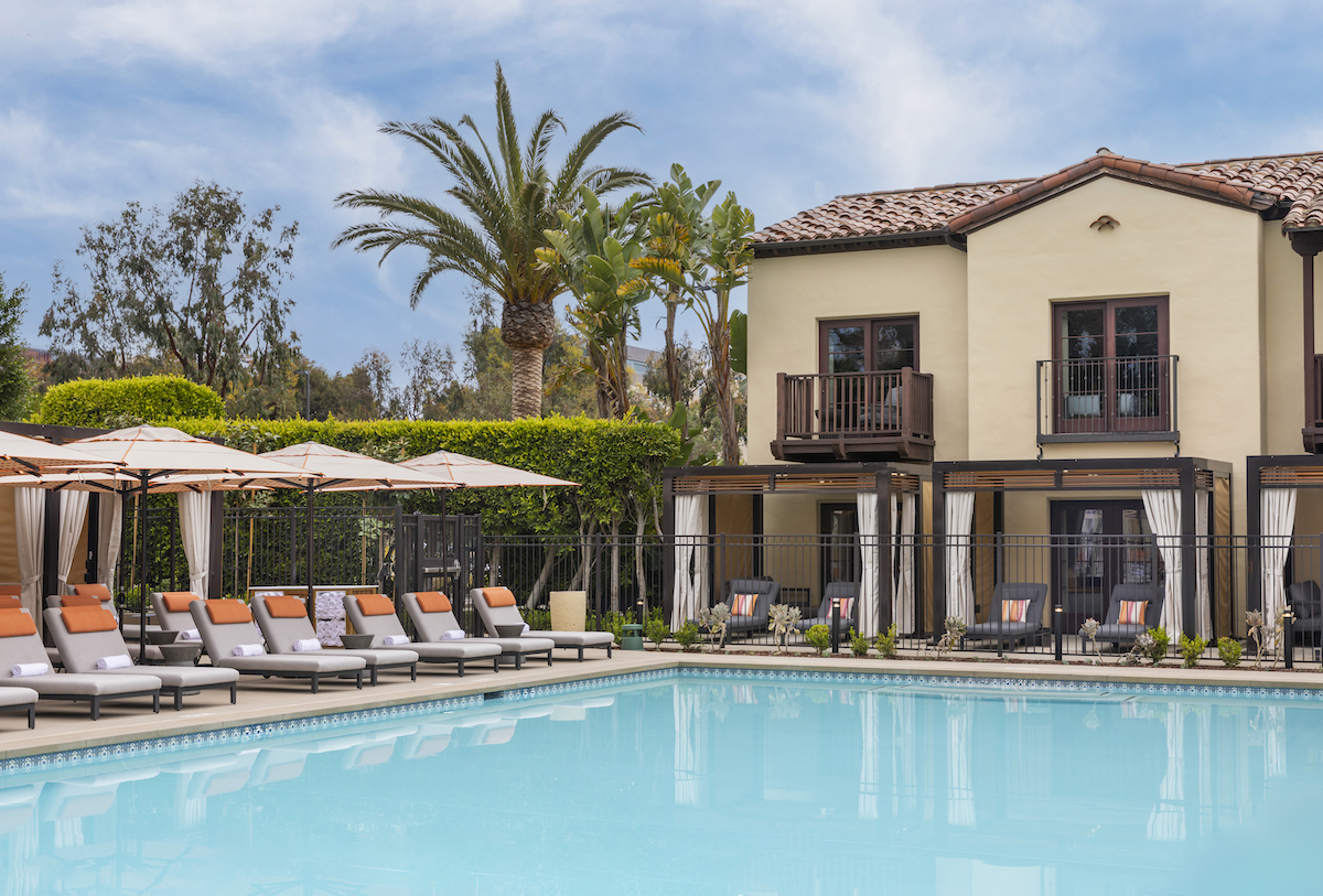 A luxurious outdoor pool area with lounge chairs and umbrellas in front of a two-story building with balconies, set against a backdrop of palm trees and hedges.