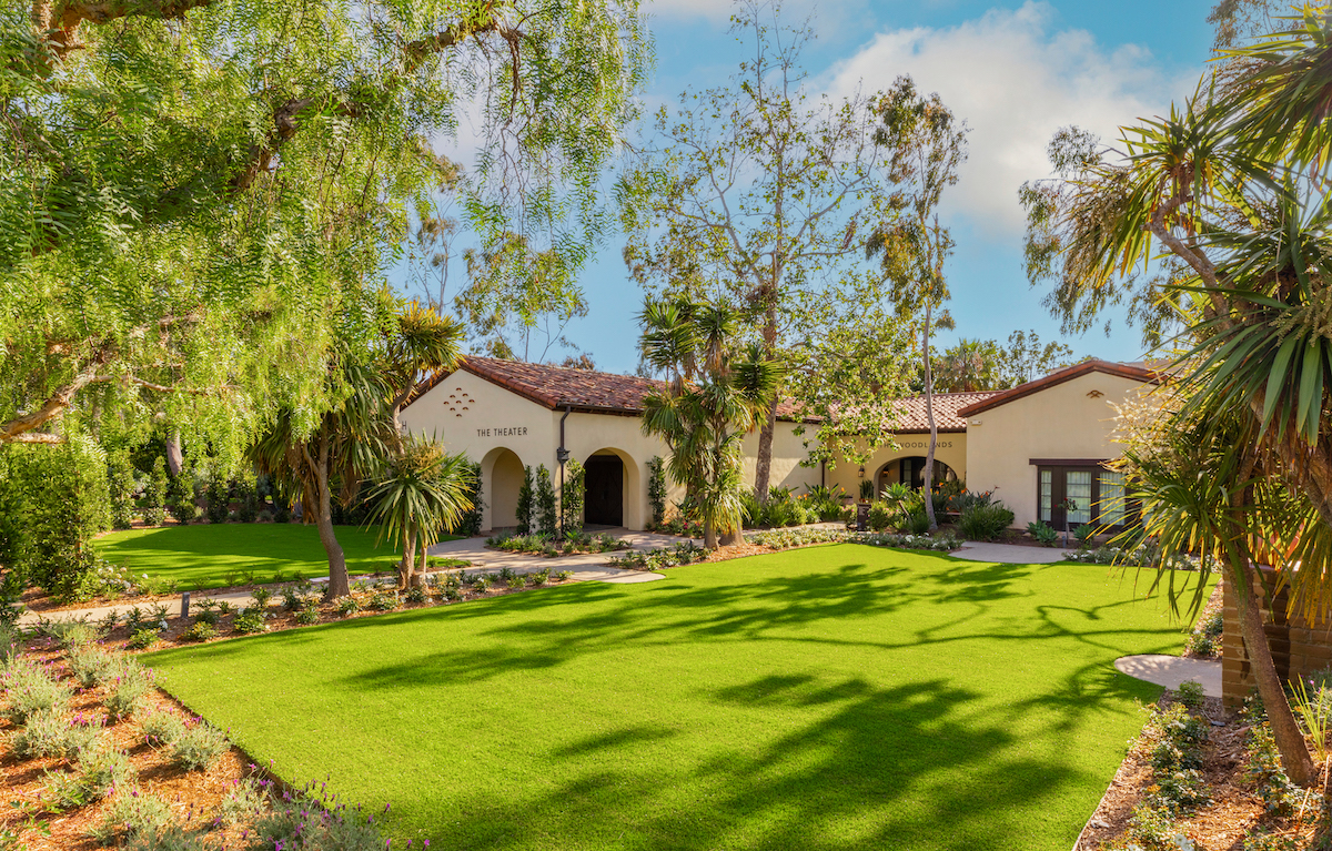 Lush garden with green lawn surrounded by trees, leading to a building labeled "Theater" in a sunny setting.