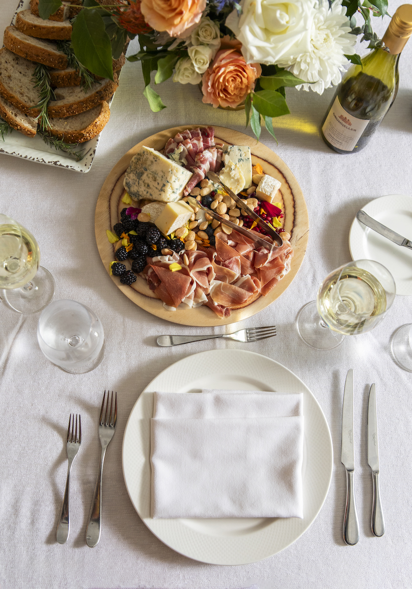 A charcuterie board with meats, cheeses, fruits, and nuts on a table set with plates, utensils, wine glasses, and a bottle of white wine. A floral arrangement and bread are in the background.