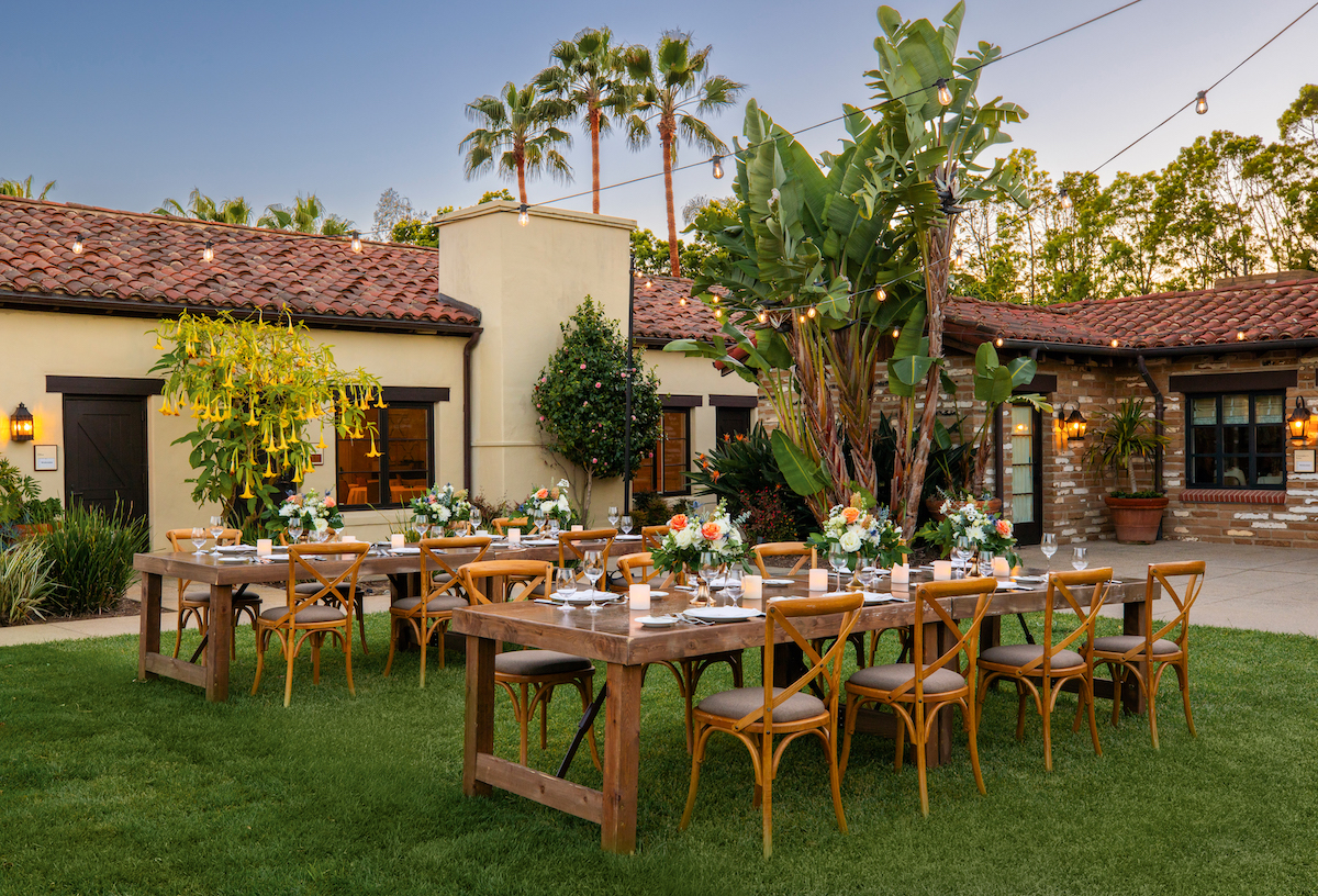 An outdoor dining setup with two wooden tables, arranged with floral centerpieces and tableware, on a grassy area near a rustic building with string lights overhead and palm trees in the background.