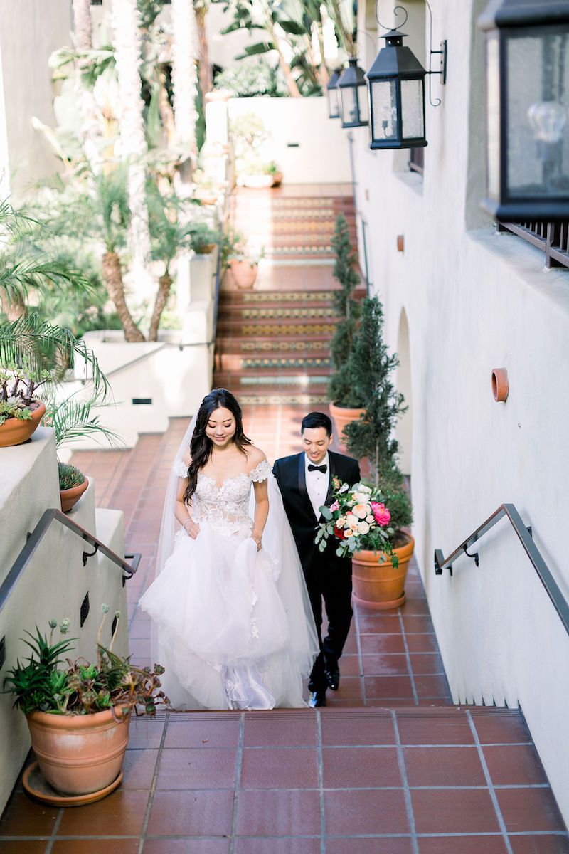 A bride in a white gown and a groom in a black suit walk up outdoor stairs, surrounded by potted plants and white walls.