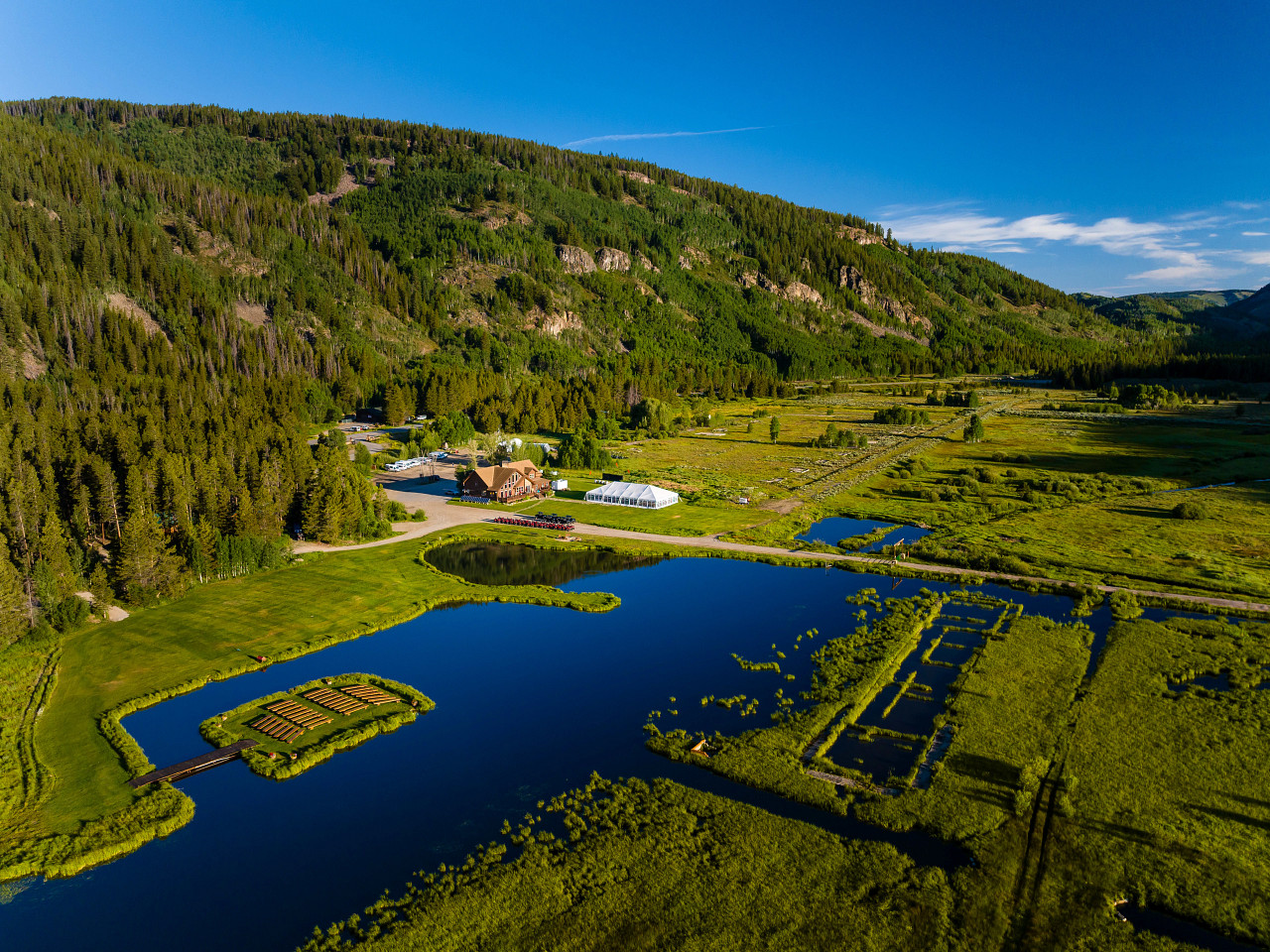 Aerial view of a large green valley with a reflective blue pond, surrounded by lush forests and hills under a clear blue sky.