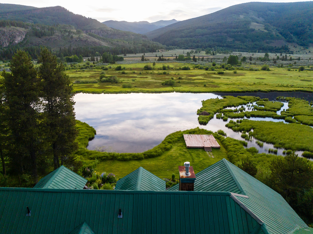 Aerial view of a house with a green roof near a pond and dock, surrounded by lush greenery and mountains in the background.