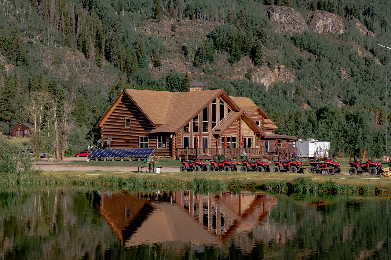 A large wooden cabin with multiple triangular windows is reflected in a pond. Several ATVs are parked in front, and solar panels sit on the ground nearby.