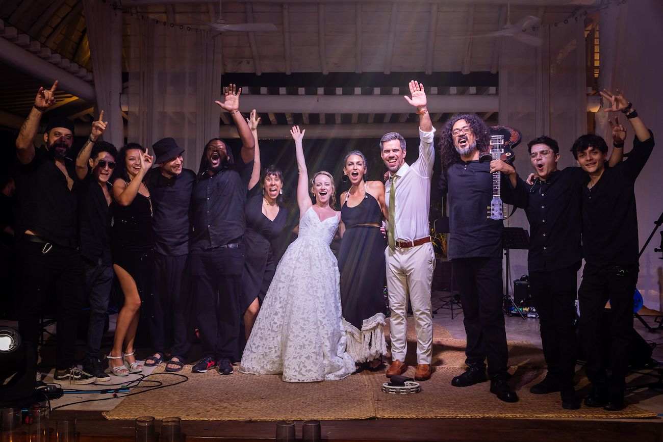 A group of people celebrates, including a bride in a white dress and a groom in a white suit, posing together with arms raised at a festive event.