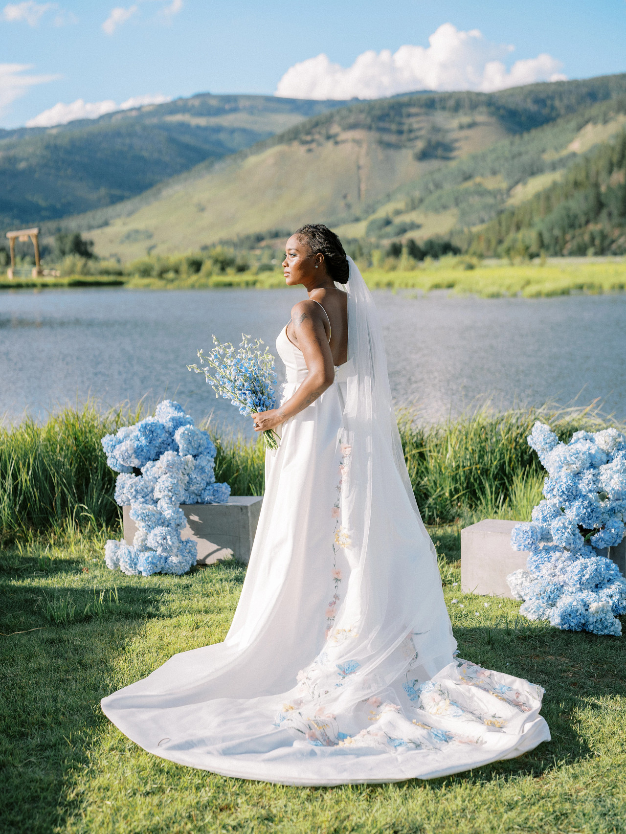 A bride in a white wedding dress and veil stands by a lakeside, holding blue flowers. Blue floral arrangements and mountains in the background.