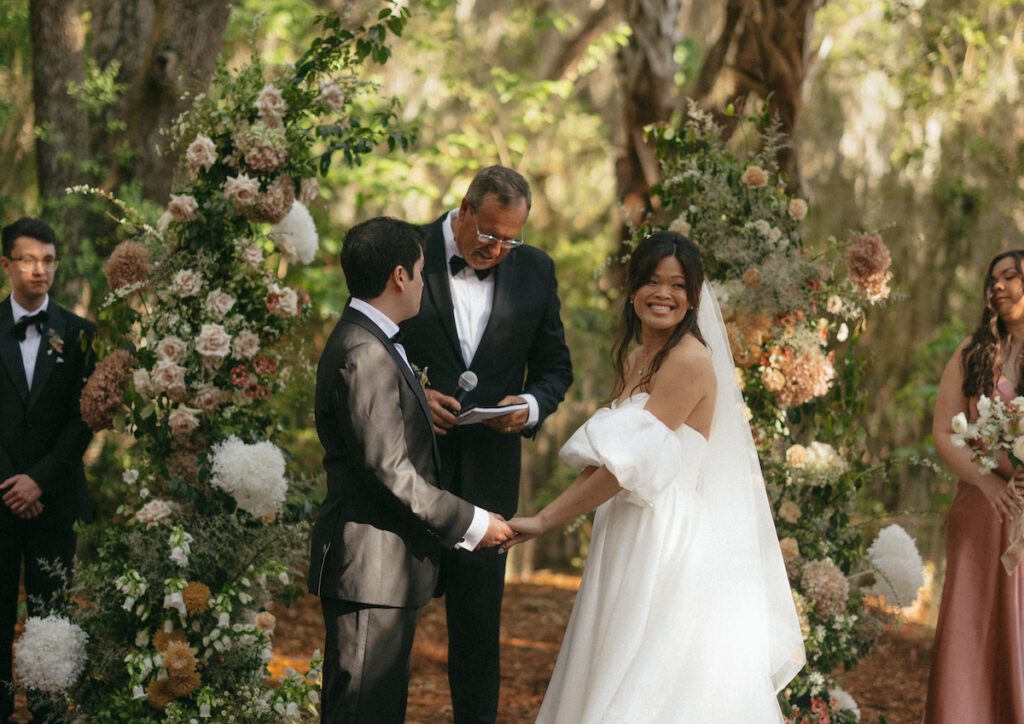 A bride and groom hold hands during an outdoor wedding ceremony, with a celebrant and wedding party standing nearby. Floral arrangements surround them.