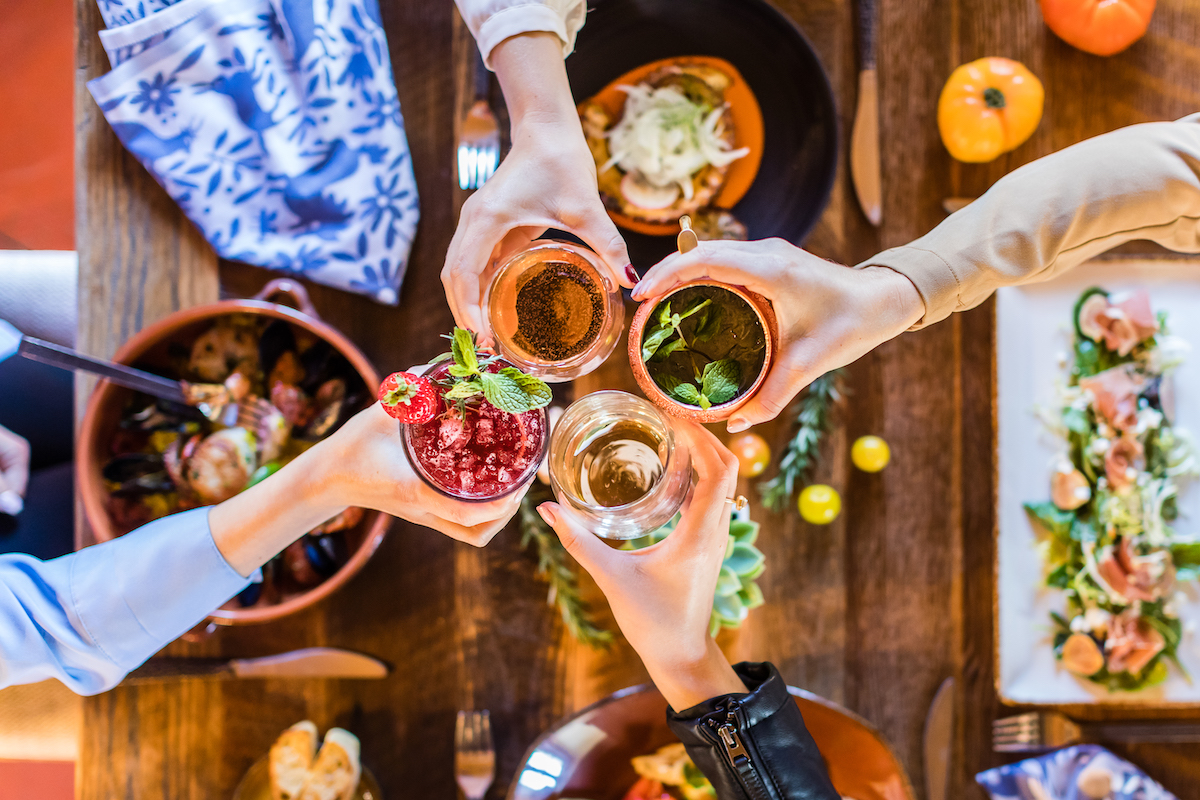 Four hands holding different cocktails over a table with assorted dishes, including a salad and hors d'oeuvres, viewed from above.