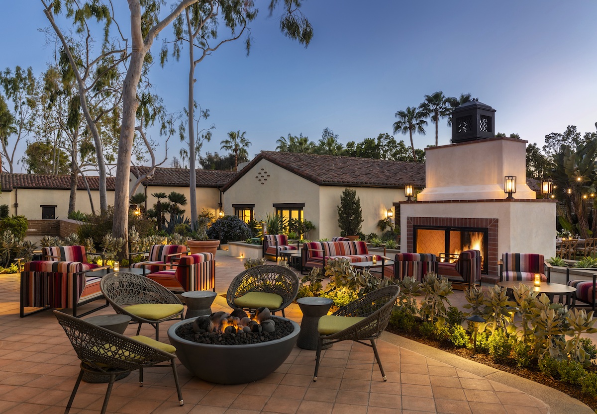 Outdoor patio with seating around a fire pit, lit fireplace, and trees. Building with red-tiled roof in the background at dusk.