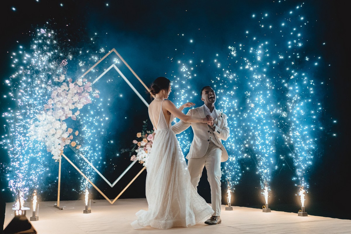 A couple dances joyfully on a stage, surrounded by sparklers and floral decorations, under a night sky.