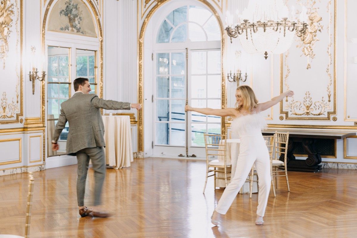 Two people in formal attire dance energetically in an elegant ballroom with chandeliers and ornate decor.